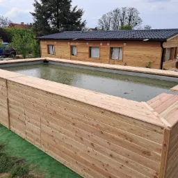 Wysoki, drewniany basen ogrodowy z szarą wodą, obok drewniany domek letniskowy z czarnym dachem, widok z góry na tle zieleni i błękitnego nieba.