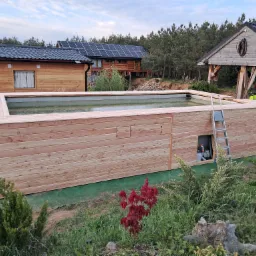 Nowoczesny, drewniany basen zewnętrzny z drabinką, otoczony zielenią, na tle zabudowań z panelami słonecznymi.