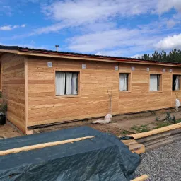 Nowoczesna, parterowa konstrukcja z jasnego drewna z oknami i czarnym dachem, otoczona zielenią i elementami budowlanymi, częściowo przykryta plandeką, w tle drzewa i błękitne niebo.