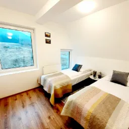 Jasny pokój z dwoma pojedynczymi łóżkami, drewnianą podłogą i widokiem na krajobraz za oknem.