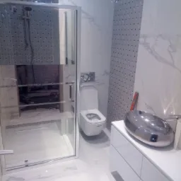 Nowoczesna łazienka z kabiną prysznicową, toaletą i umywalką w kształcie misy, wykończona białymi marmurkowymi płytkami i mozaiką w odcieniach szarości.