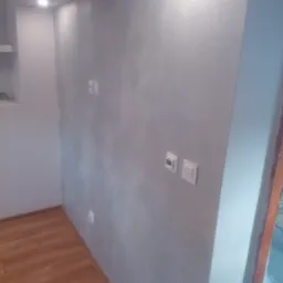 Ściana w pokoju po nałożeniu dekoracyjnej, srebrzystej farby z efektem perłowym, widoczne gniazdka elektryczne i drewniana podłoga.