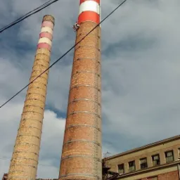 Pracownik na wysokości podczas malowania komina przemysłowego w biało-czerwone pasy; drugi, starszy komin w tle, budynek przemysłowy z cegły po prawej.