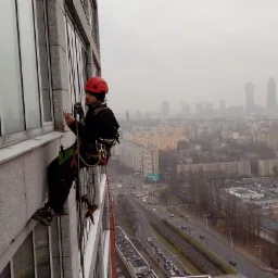 Pracownik w czerwonym kasku, zabezpieczony linami, w trakcie pracy na wysokości przy szklanej elewacji biurowca, z widokiem na miasto w tle.