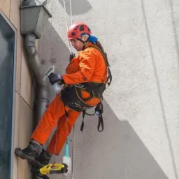 Pracownik w pomarańczowym kombinezonie i czerwonym kasku, zawieszony na linach, spryskuje i czyści okno budynku. Widoczny fragment elewacji, rynna i drabinka linowa.