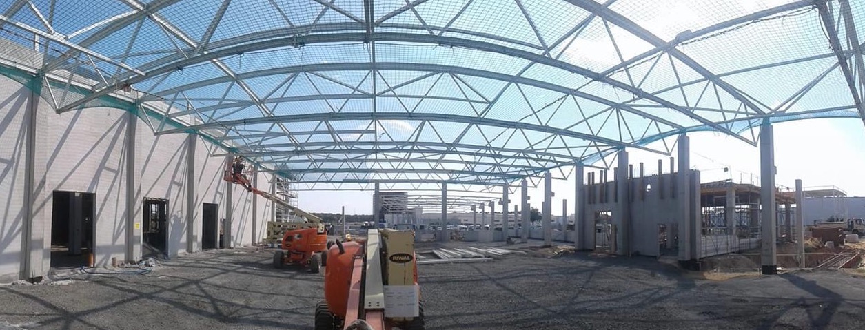 Wnętrze hali w trakcie budowy z widoczną konstrukcją dachu zabezpieczoną siatką, betonowymi słupami i pracownikiem na podnośniku koszowym.