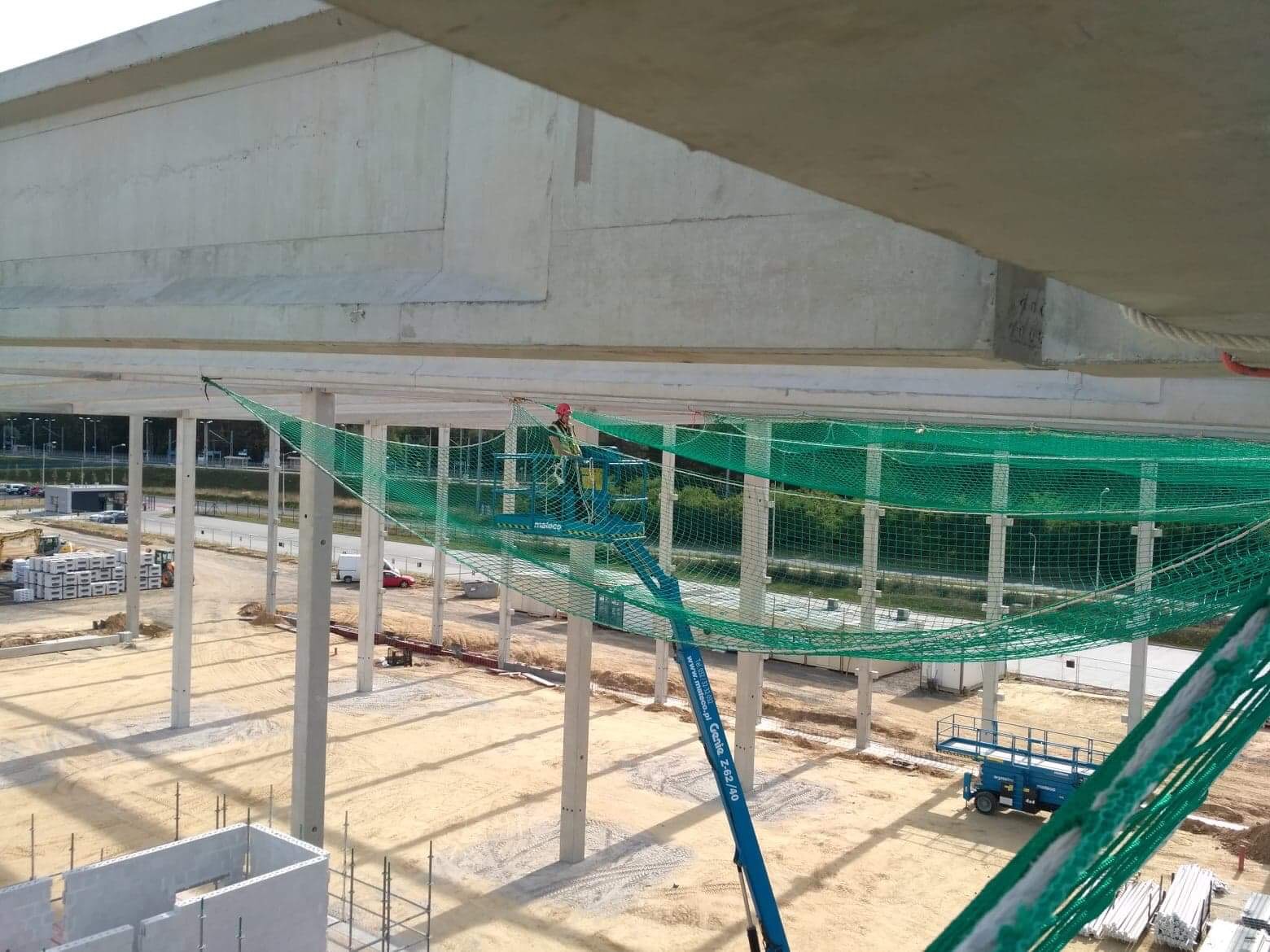 Pracownik w czerwonym kasku na podnośniku koszowym, montujący siatkę zabezpieczającą pod betonowym stropem hali w trakcie budowy.