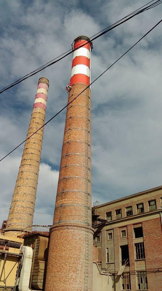 Pracownik na wysokości podczas malowania komina przemysłowego w biało-czerwone pasy; drugi, starszy komin w tle, budynek przemysłowy z cegły po prawej.