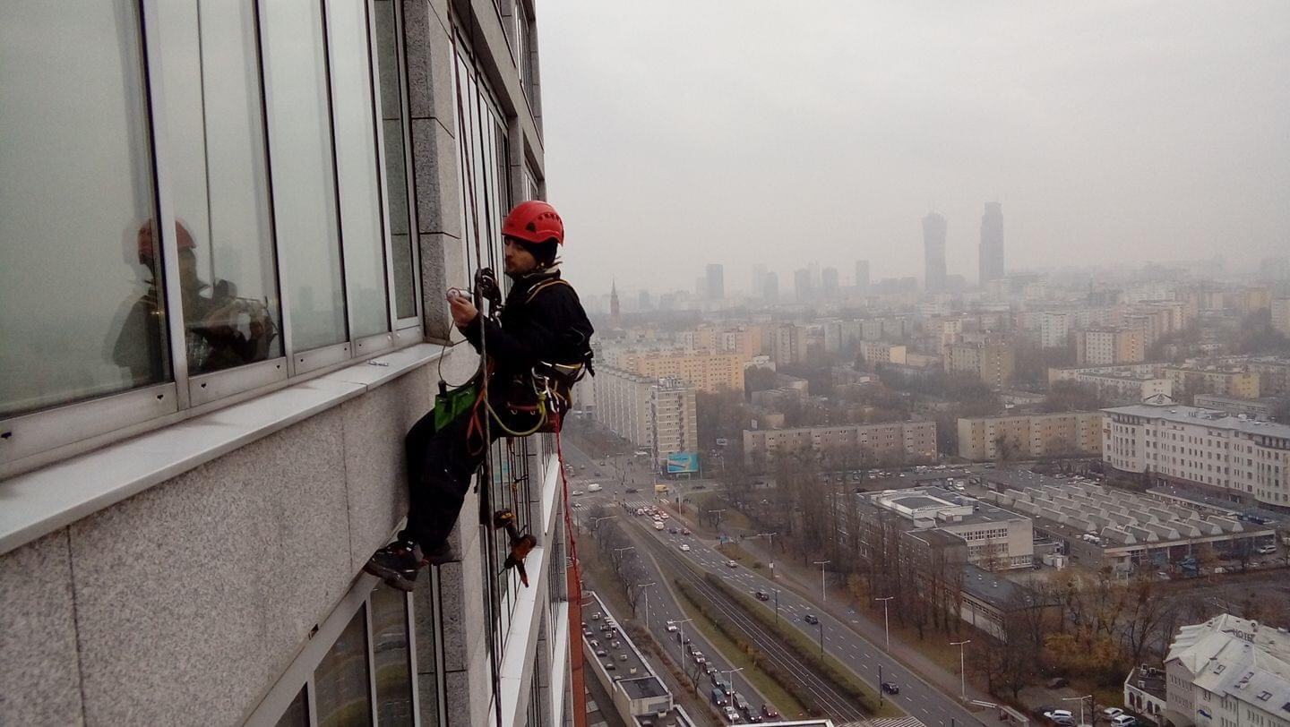 Pracownik w czerwonym kasku, zabezpieczony linami, w trakcie pracy na wysokości przy szklanej elewacji biurowca, z widokiem na miasto w tle.