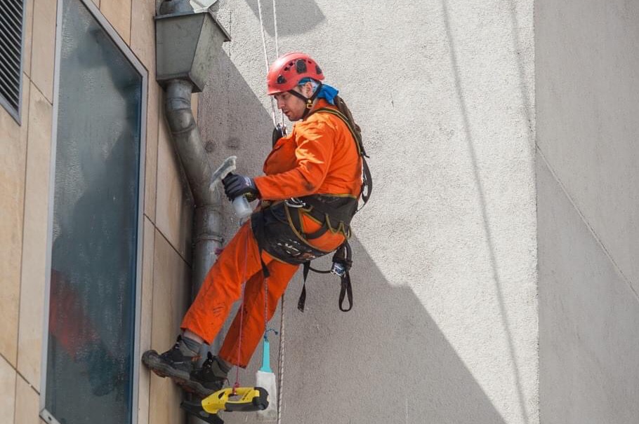 Pracownik w pomarańczowym kombinezonie i czerwonym kasku, zawieszony na linach, spryskuje i czyści okno budynku. Widoczny fragment elewacji, rynna i drabinka linowa.