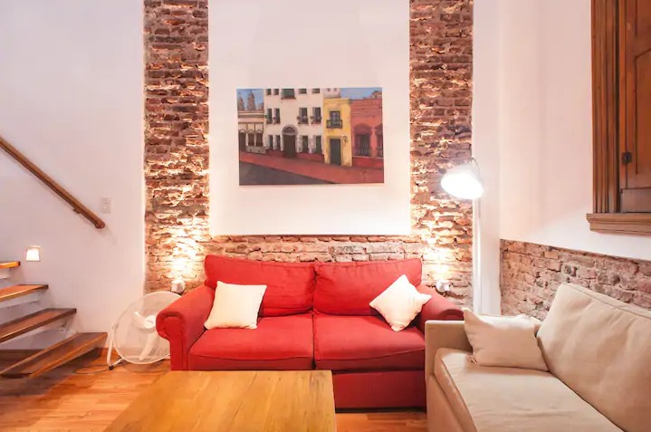 Ceglana ściana z obrazem w salonie, czerwona sofa z białymi poduszkami, drewniane schody i parkiet, wentylator pod schodami.