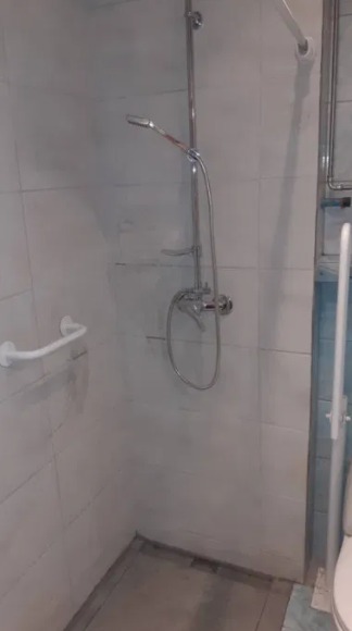 Biała łazienka z zamontowanym chromowanym prysznicem, uchwytem dla niepełnosprawnych i widoczną częścią toalety.