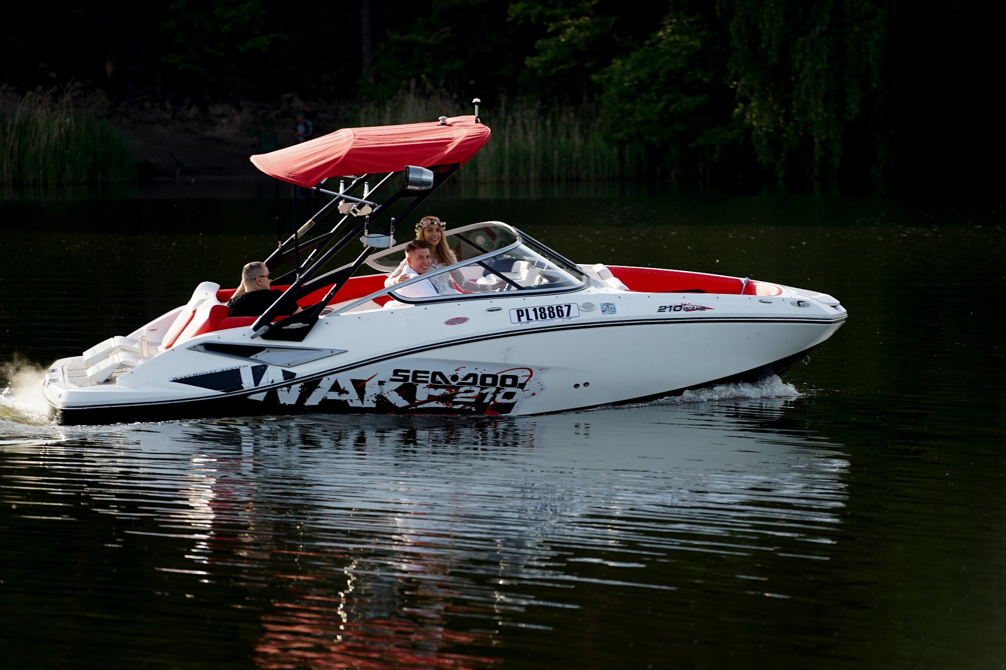 Młoda para w białej łodzi motorowej SeaDoo 210 Wake z czerwonymi akcentami i baldachimem, płynąca po jeziorze, widoczny numer rejestracyjny PL18867.