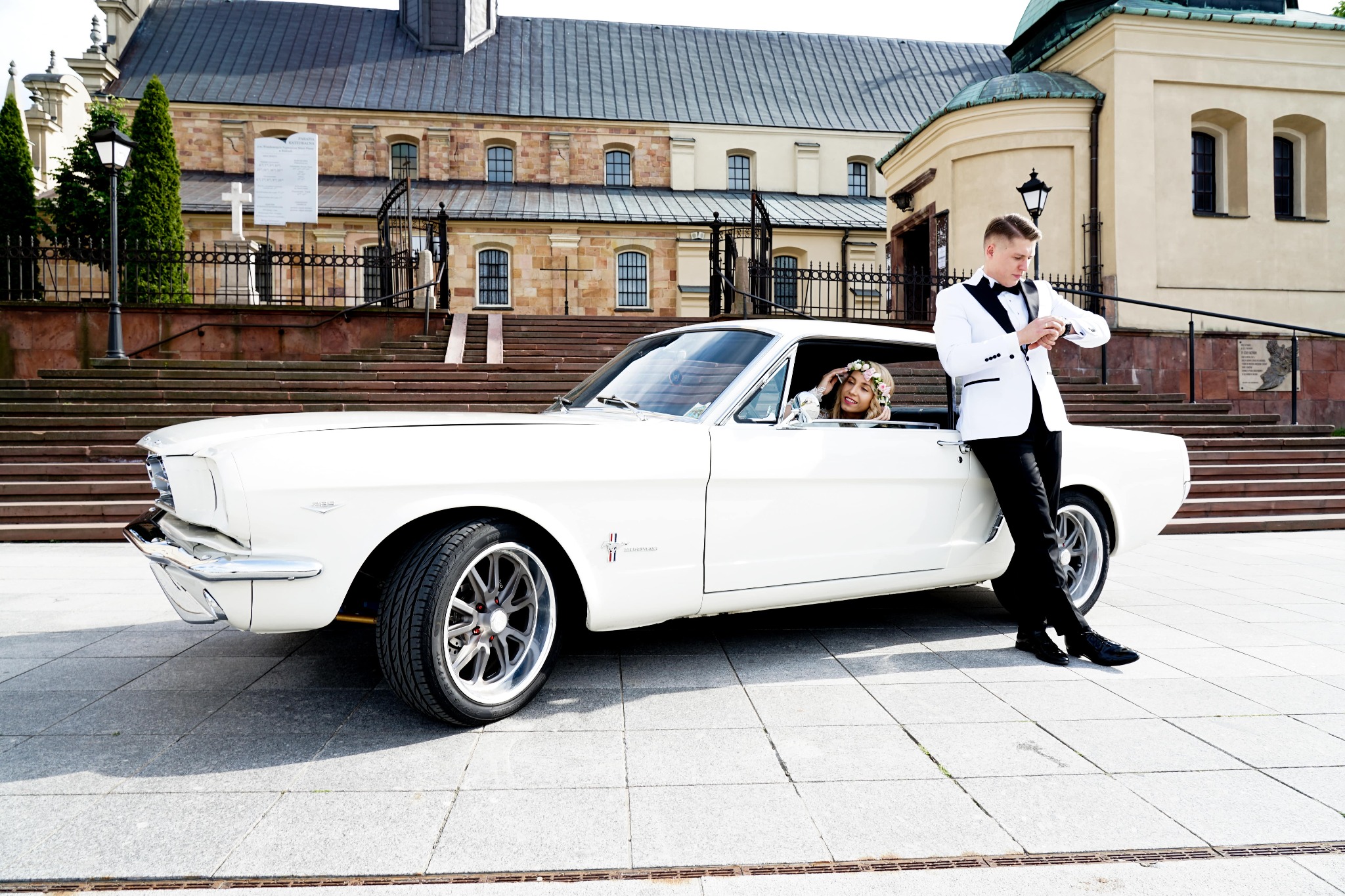 Elegancki mężczyzna w białym smokingu sprawdza godzinę przy białym klasycznym samochodzie, w którym siedzi uśmiechnięta kobieta z wiankiem na głowie, na tle budynku z cegły.
