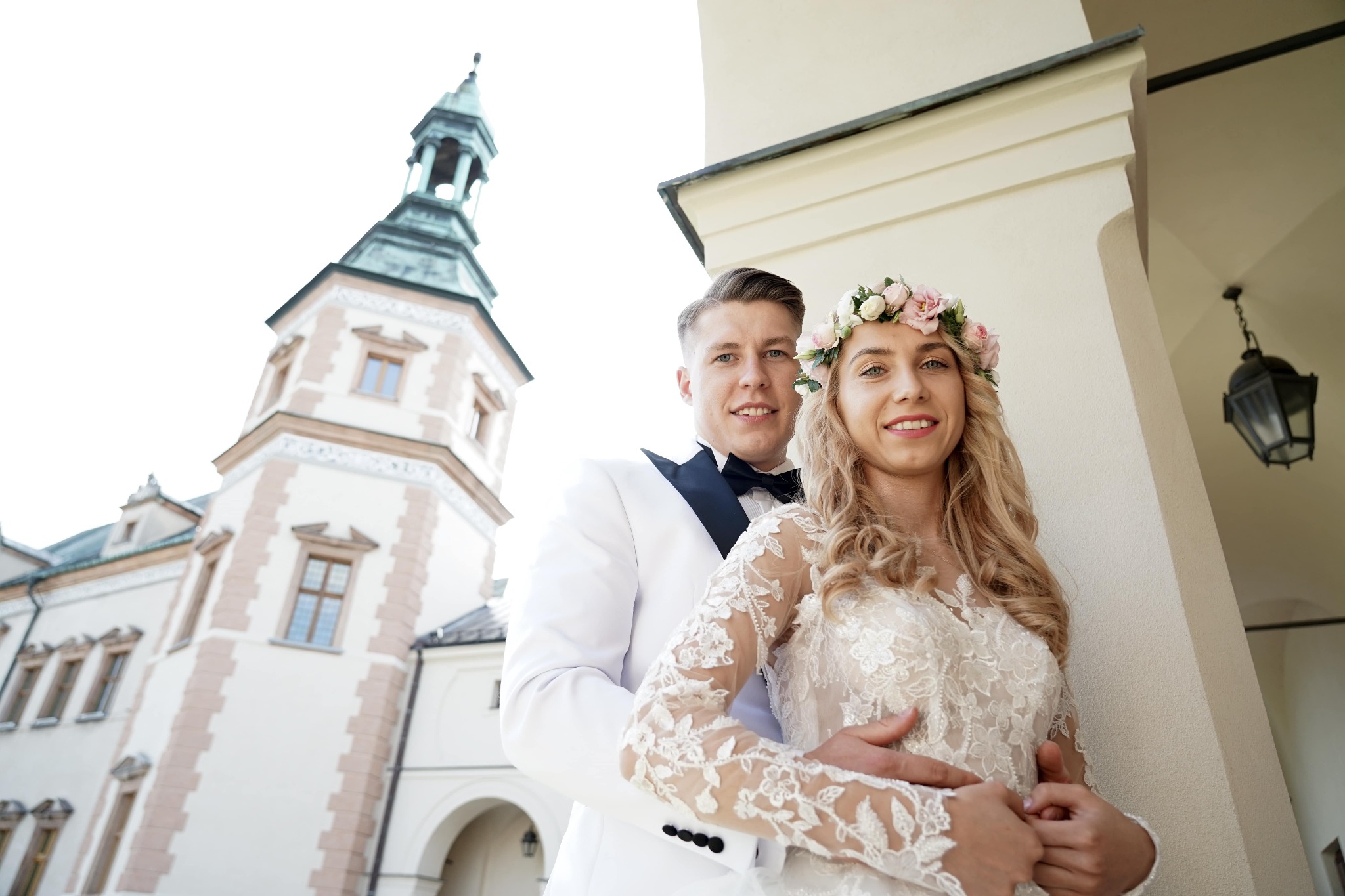 Uśmiechnięta para młoda: pan młody w białym smokingu obejmuje pannę młodą w koronkowej sukni i wianku na tle wieży zamkowej.
