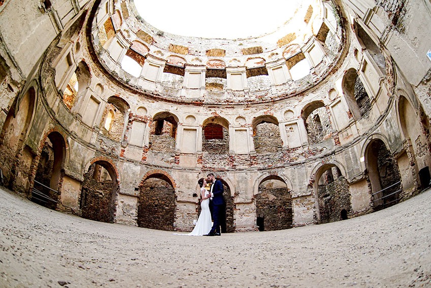 Para młoda całuje się w ruinach okrągłego zamku z cegły, z otwartym dachem i licznymi arkadami.
