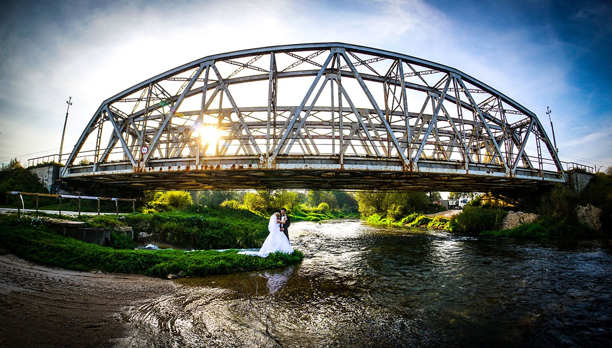 Para młoda w strojach ślubnych stoi na brzegu rzeki pod metalowym mostem w słoneczny dzień.