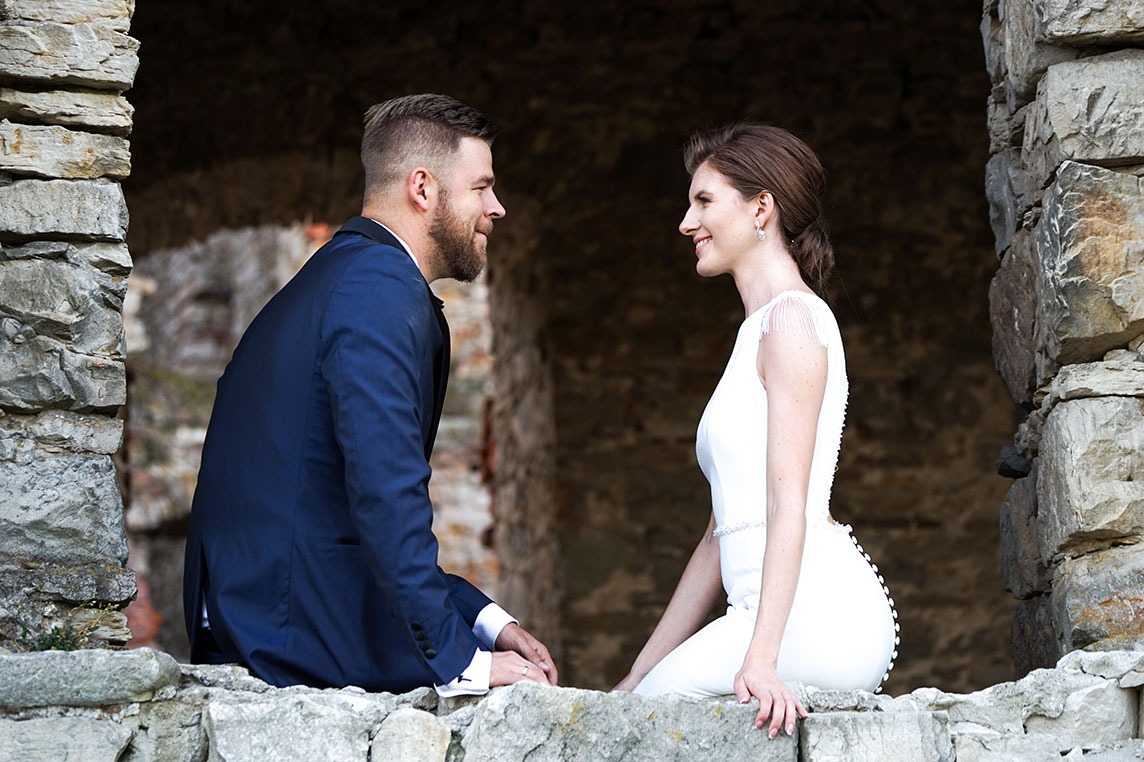 Uśmiechnięta para młoda siedzi na kamiennym murze, on w granatowym garniturze, ona w prostej białej sukni, patrzą na siebie z czułością.