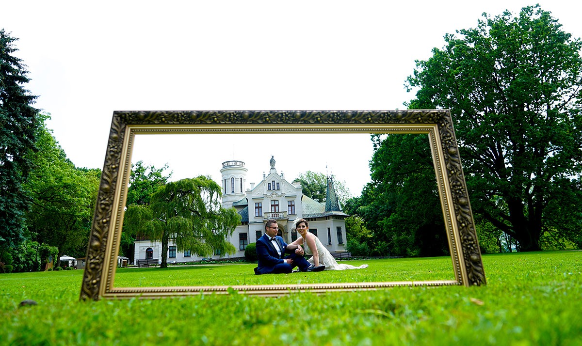 Para młoda siedząca na trawie, ujęta w ozdobną ramę z widokiem na pałac w tle.