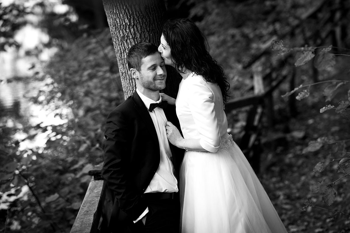 Czarno-białe zdjęcie pary młodej: panna młoda w sukni ślubnej całuje pana młodego w czoło, opierającego się o drzewo na tle leśnej scenerii.
