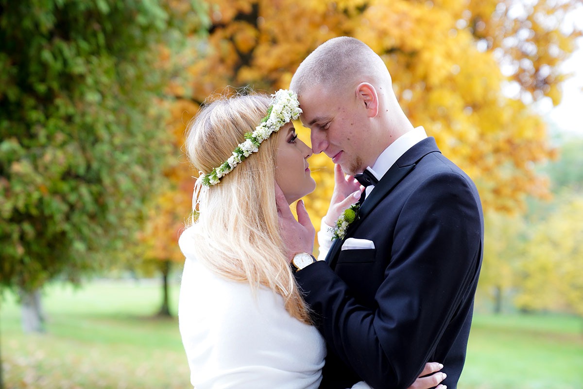 Para młoda w jesiennej scenerii: panna młoda w wianku z białych kwiatów i narzeczony w czarnym garniturze z muchą, delikatny dotyk dłoni i spojrzenia.