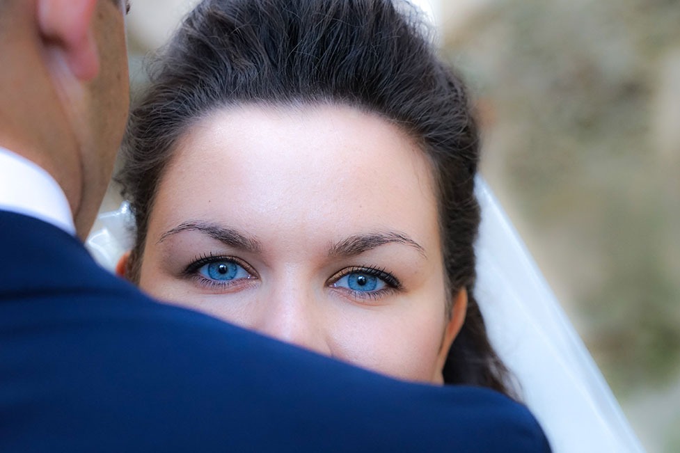 Zbliżenie na oczy panny młodej o intensywnym, niebieskim kolorze, spoglądającej zza ramienia pana młodego w granatowym garniturze, z delikatnym welonem w tle.