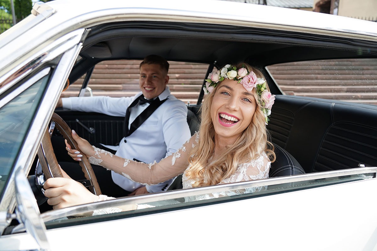 Uśmiechnięta panna młoda w wianku z różowych kwiatów siedzi za kierownicą białego, klasycznego samochodu, obok niej widać pana młodego w białym smokingu.