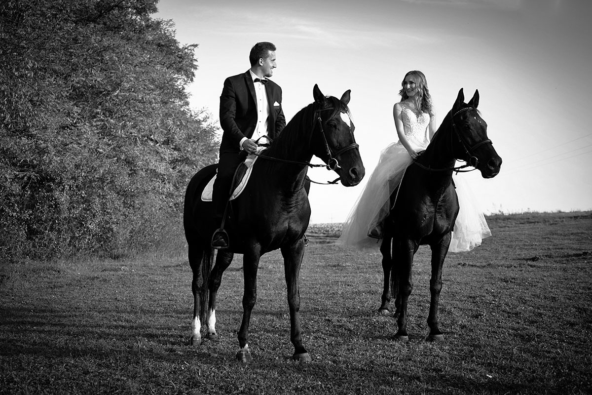 Czarno-białe zdjęcie pary młodej na koniach na łące, pan młody w smokingu, panna młoda w sukni ślubnej z tiulową spódnicą, oboje uśmiechnięci, zarys drzew w tle.