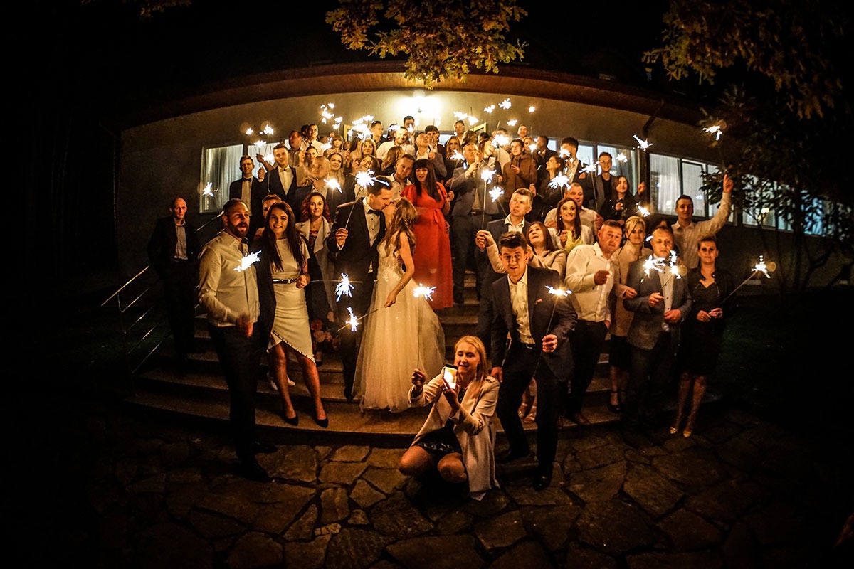 Grupa gości weselnych z zimnymi ogniami na schodach przed budynkiem, para młoda w centrum, kobieta robiąca zdjęcie telefonem na pierwszym planie, nocne oświetlenie.