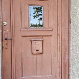 Drew-klas - Drewniane drzwi wejściowe w kolorze brązowym, z małym okienkiem i skrzynką na listy, widoczne ślady użytkowania i odpryski farby.
