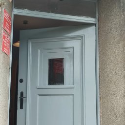 Drew-klas - Pomalowane na szaro drzwi wejściowe z małym okienkiem, widoczne od zewnątrz, z tabliczką informacyjną po lewej stronie, nad drzwiami lampa.