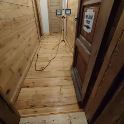 Wąski korytarz wyłożony drewnianymi panelami ściennymi i podłogowymi, oświetlony dwoma lampami halogenowymi na statywie, z widocznym napisem 'Wejście Obok' na drzwiach po prawej stronie.