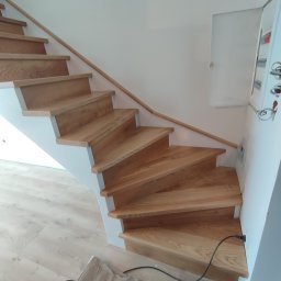 Drew-klas - Jasne drewniane schody z białymi podstopnicami i drewnianą poręczą, widok z dołu, kabel zasilający wystający ze ściany przy schodach.