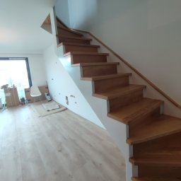 Drew-klas - Nowoczesne schody drewniane z jasnego drewna, z białymi podstopnicami i prostą drewnianą balustradą, prowadzące na piętro w jasnym wnętrzu. Widoczne niedokończone prace wykończeniowe w tle.