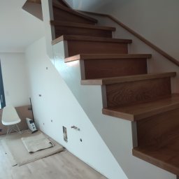 Drew-klas - Drewniane schody z białymi podstopnicami podczas montażu, widoczne przygotowania do instalacji elektrycznej w ścianie, zabezpieczona podłoga i krzesło w tle.