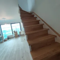 Drewniane schody z jasnymi podstopnicami i poręczą, widok z dołu, w tle okno z widokiem na budowę.