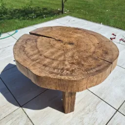 Stolik kawowy z grubego pnia drzewa z widocznymi słojami, umieszczony na tarasie z jasnych płytek, w tle trawnik i drzewo.