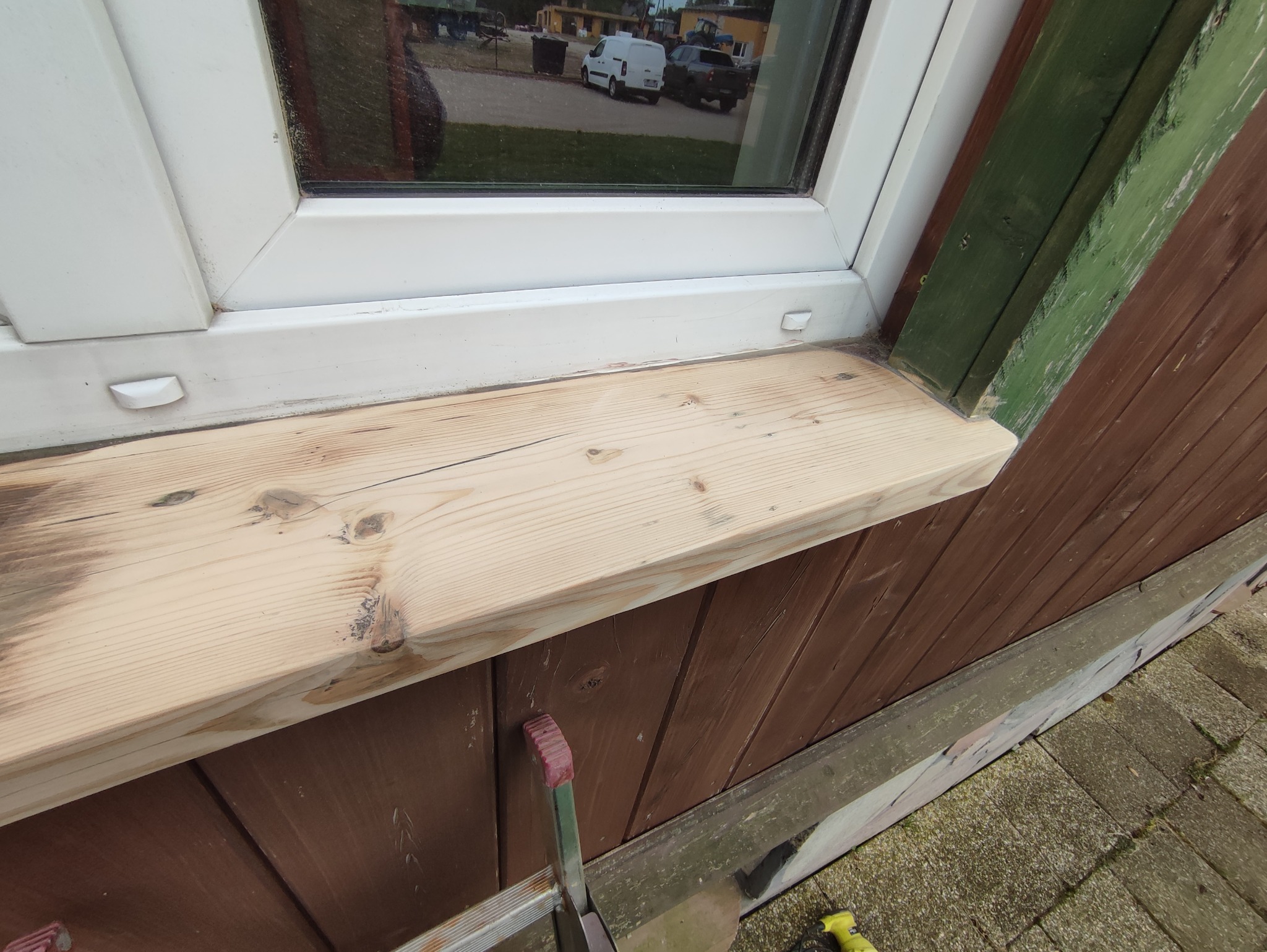 Montaż drewnianego parapetu zewnętrznego pod oknem PCV, widoczne niedomalowane elementy elewacji i drabina.