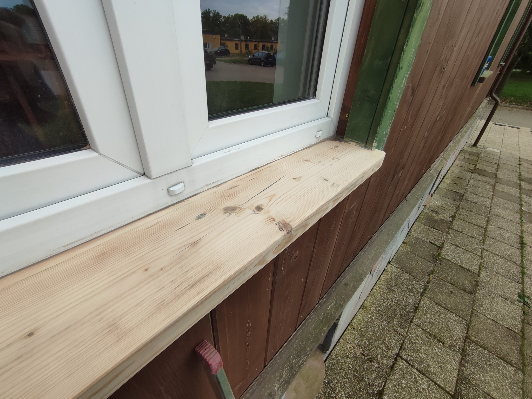 Surowe drewno parapetu zewnętrznego pod oknem PCV, widoczne słoje i detale drewna, brązowa drewniana elewacja budynku, chodnik z kostki brukowej.