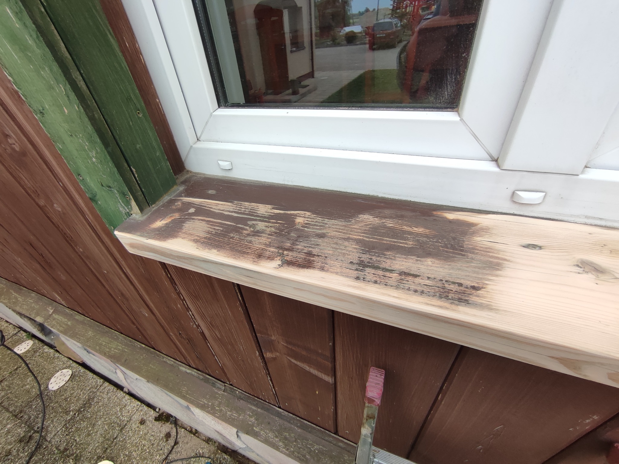 Drewniany parapet zewnętrzny w trakcie renowacji, częściowo pomalowany na brązowo, widoczny pędzel i elewacja domu z drewnianą boazerią.