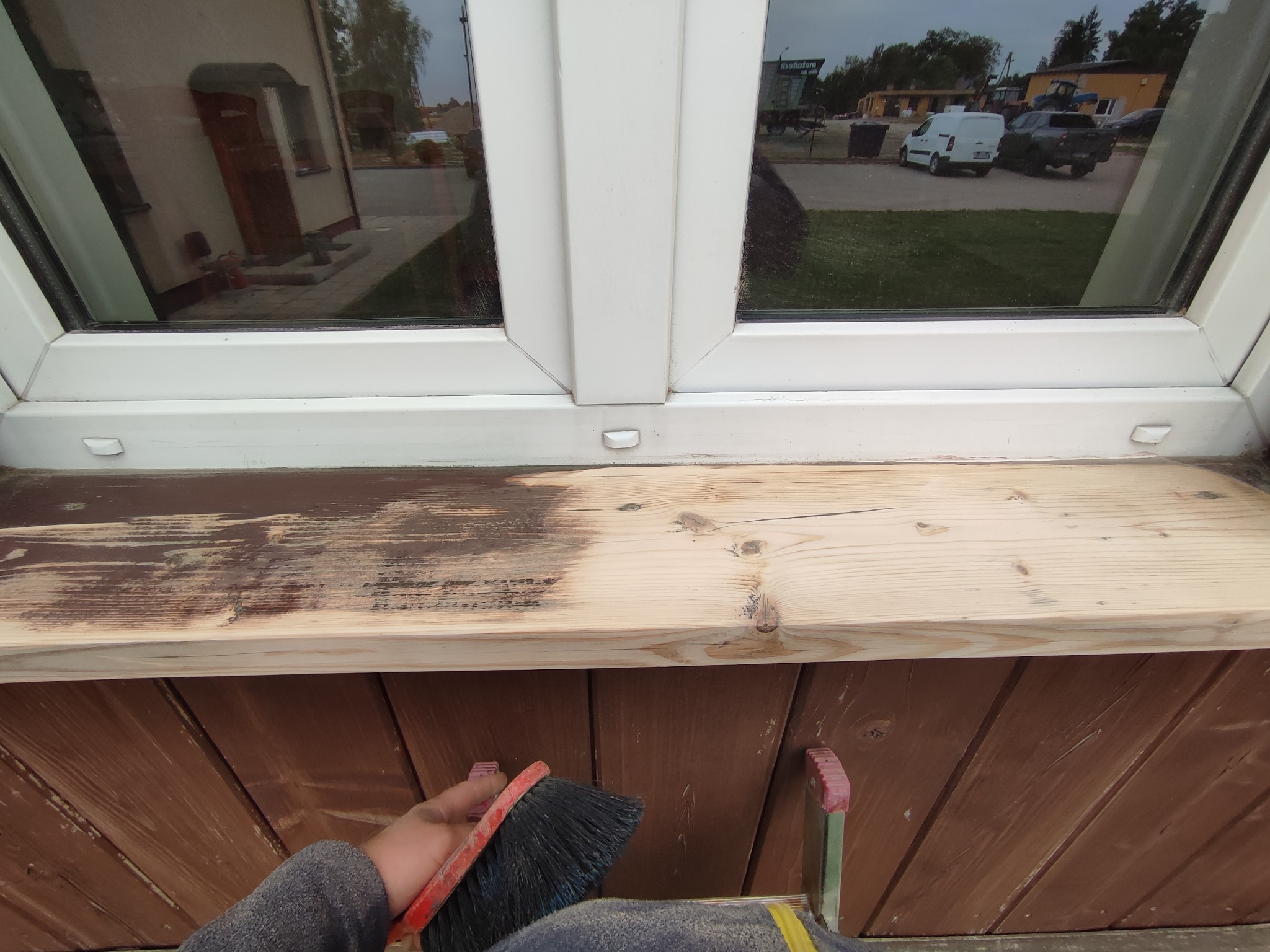 Malowanie drewnianego parapetu wewnętrznego przy oknie, widoczny pędzel i częściowo pomalowana powierzchnia, w tle odbicie otoczenia w szybie.
