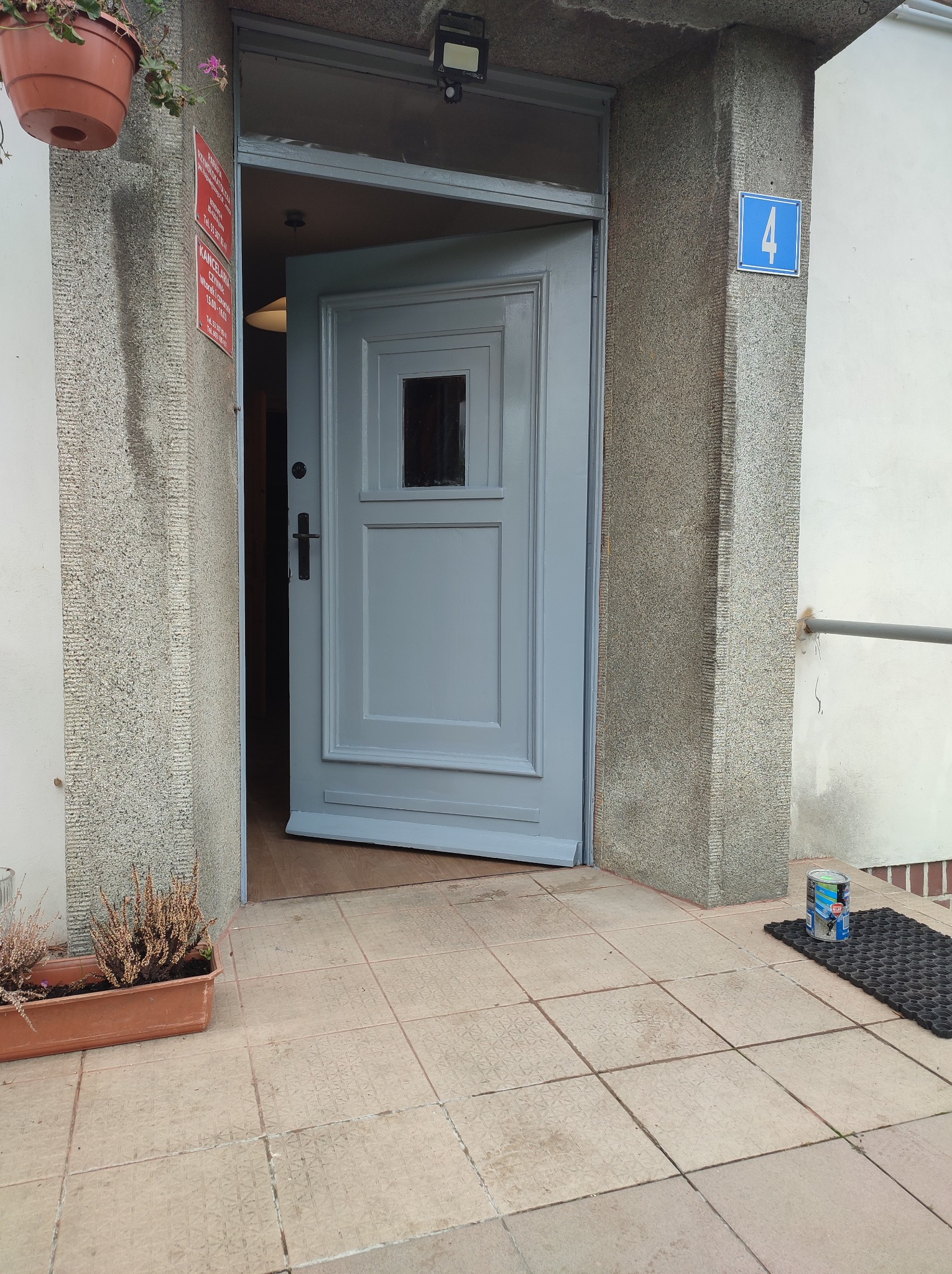 Wejście do budynku w Kmiecinie z otwartymi, szarymi drzwiami; widoczny numer '4' na ścianie, doniczki z kwiatami i puszka farby na wycieraczce.