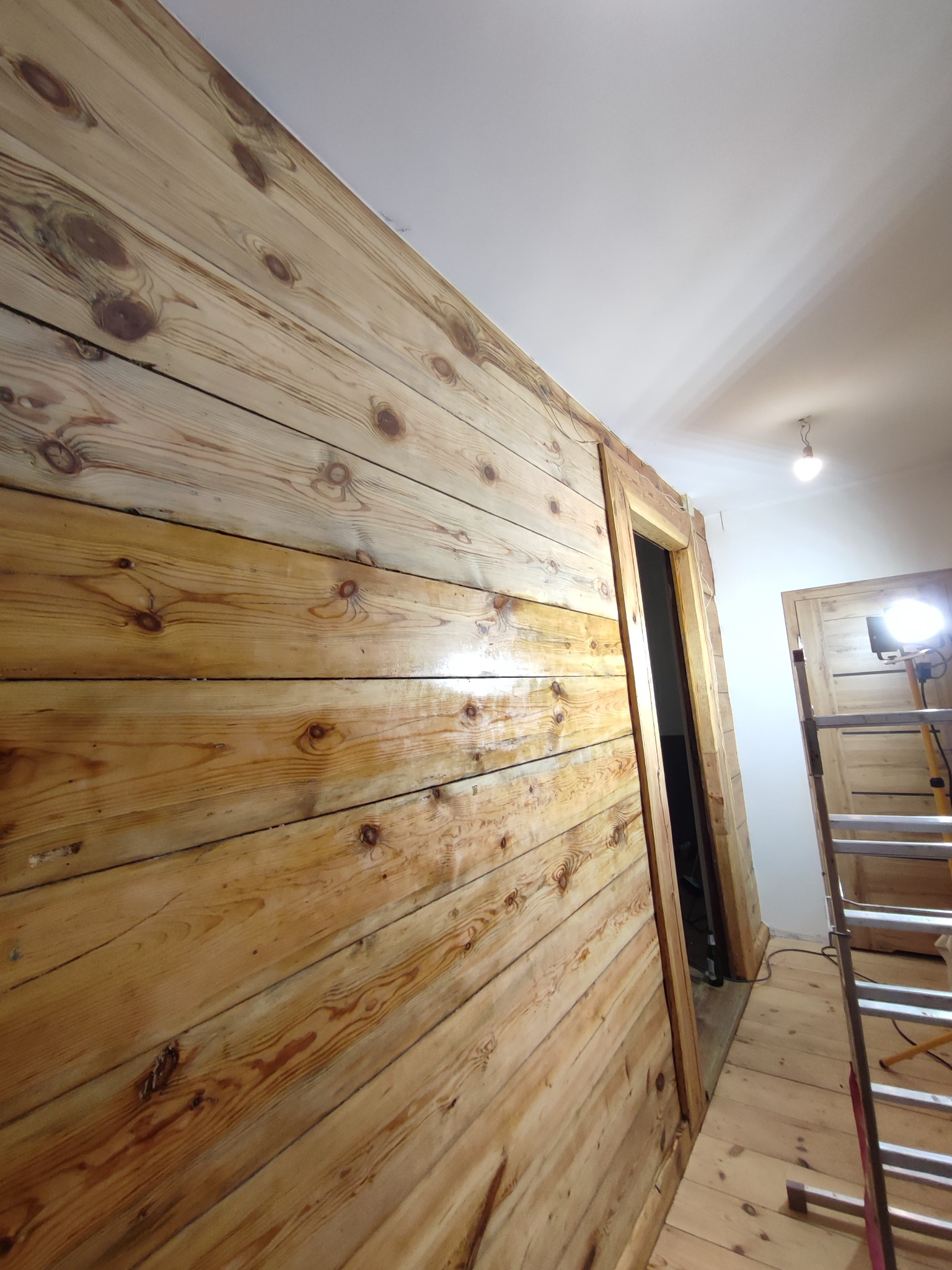 Ściana wyłożona surowymi, lakierowanymi deskami z widocznymi sękami, obok ościeżnica drzwiowa i drabina warsztatowa w słabo oświetlonym wnętrzu.