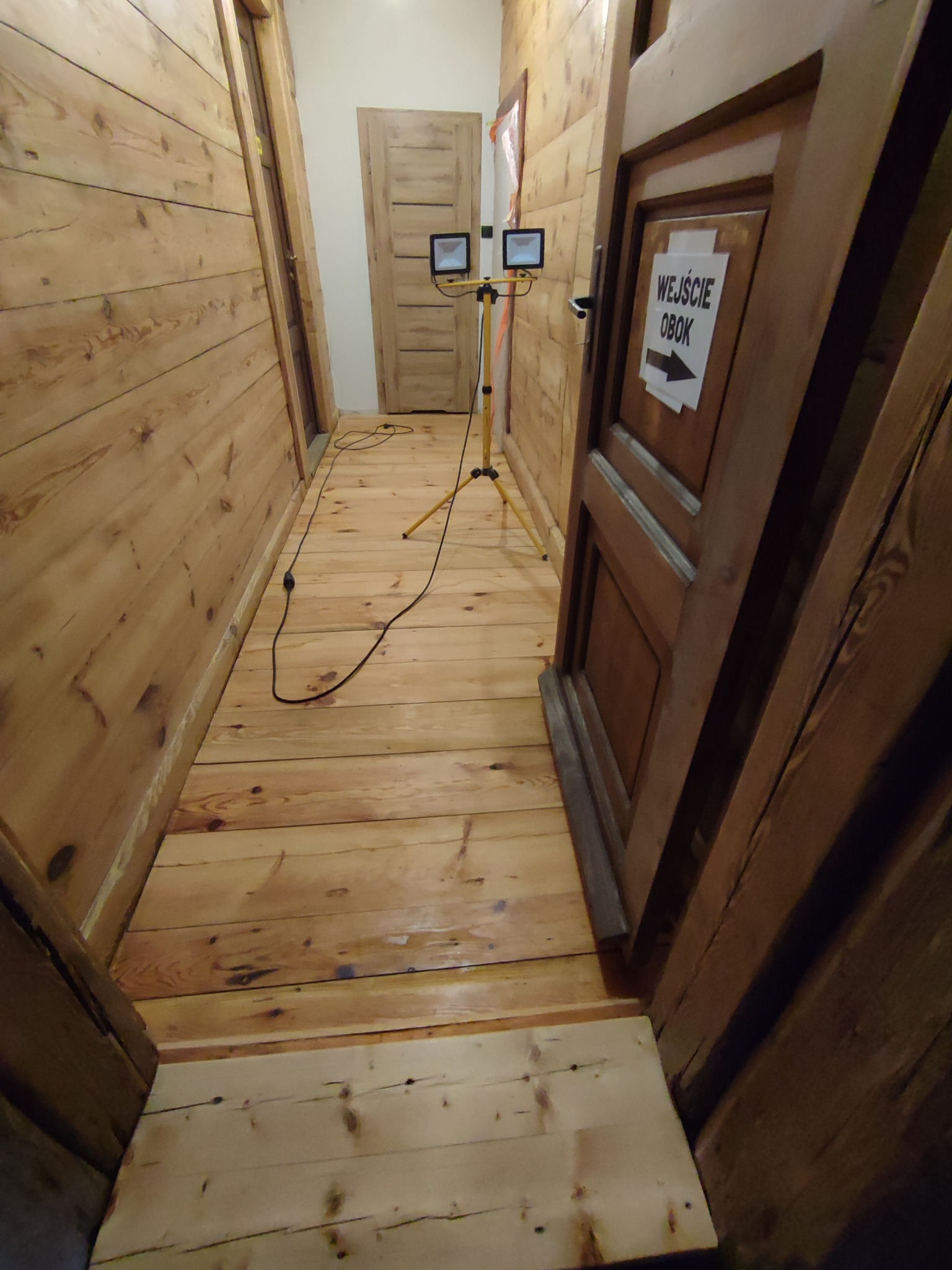 Wąski korytarz wyłożony drewnianymi panelami ściennymi i podłogowymi, oświetlony dwoma lampami halogenowymi na statywie, z widocznym napisem 'Wejście Obok' na drzwiach po prawej stronie.