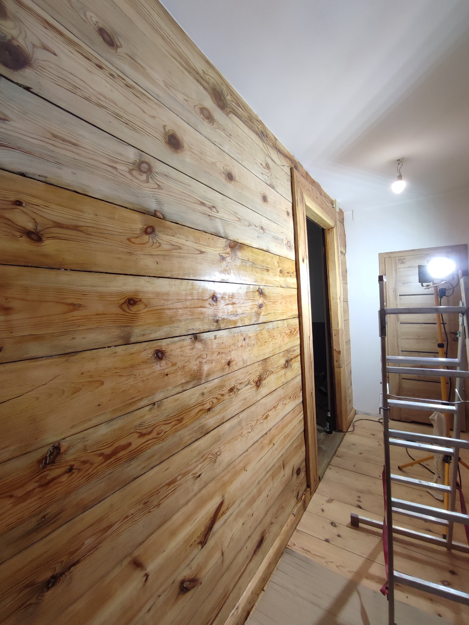 Ściana i podłoga wyłożone jasnym drewnem z widocznymi sękami, częściowo polakierowane, z drabiną i lampą budowlaną w tle, w trakcie prac wykończeniowych.