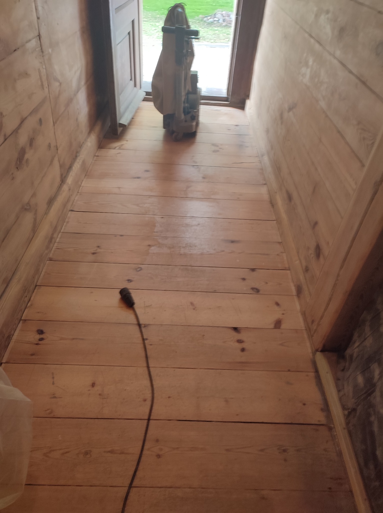 Wąski korytarz z drewnianą podłogą w trakcie renowacji, widoczna szlifierka do parkietu i ślady po szlifowaniu, drewniane ściany i drzwi w tle.