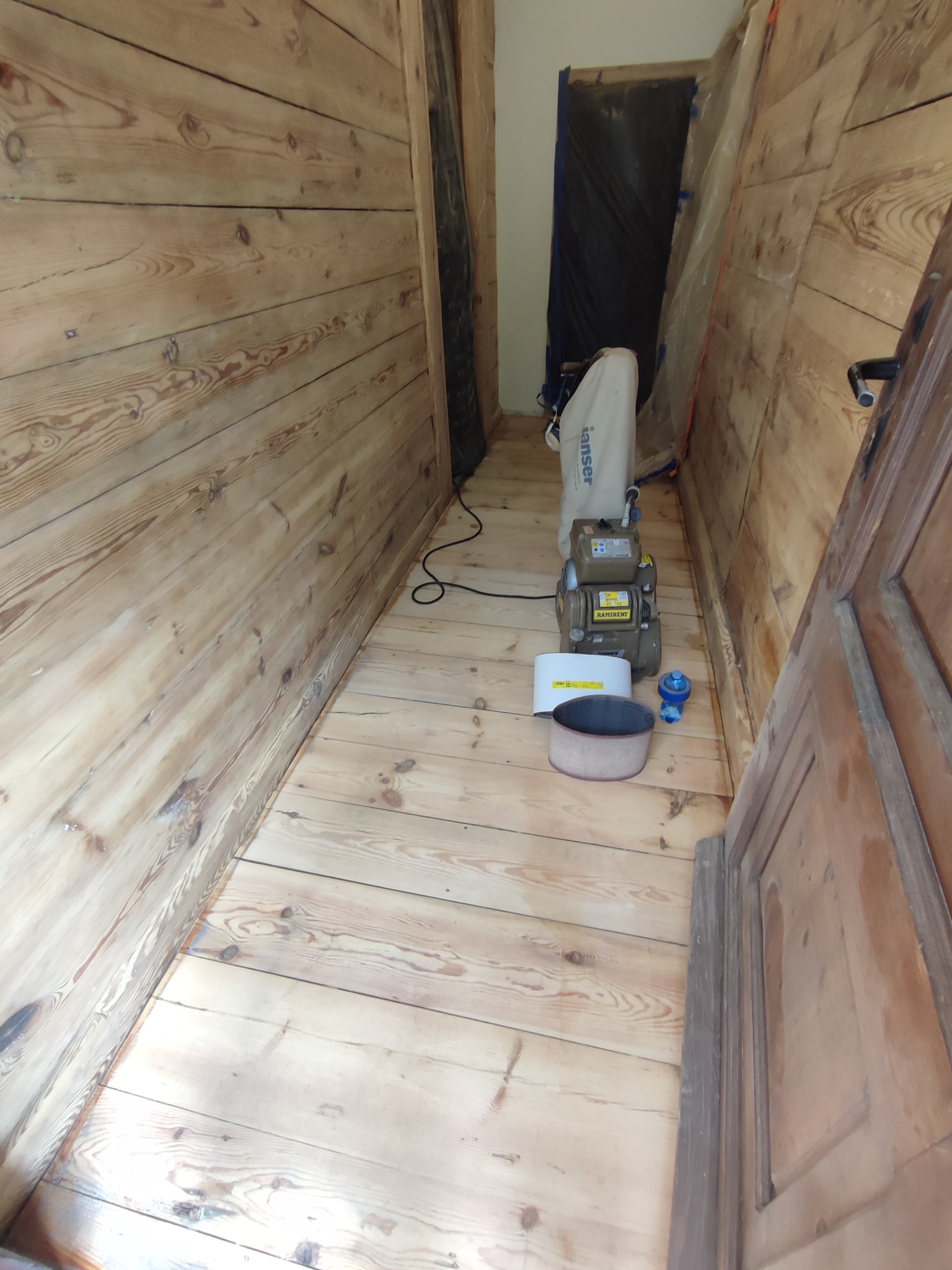 Szlifowanie drewnianej podłogi w wąskim korytarzu, widoczna maszyna do cyklinowania, ściany wyłożone boazerią, zabezpieczone drzwi folią.