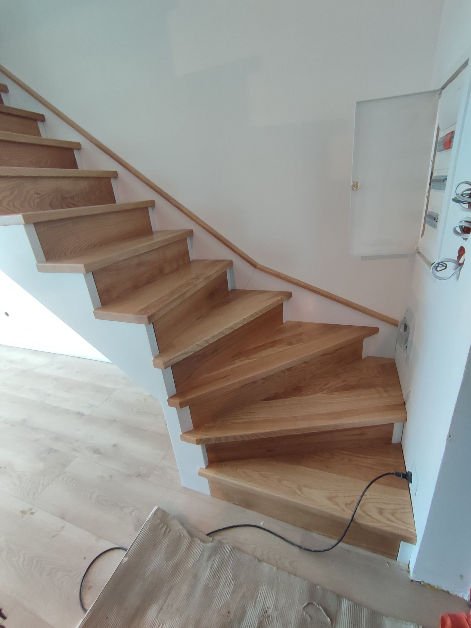 Jasne drewniane schody z białymi podstopnicami i drewnianą poręczą, widok z dołu, kabel zasilający wystający ze ściany przy schodach.