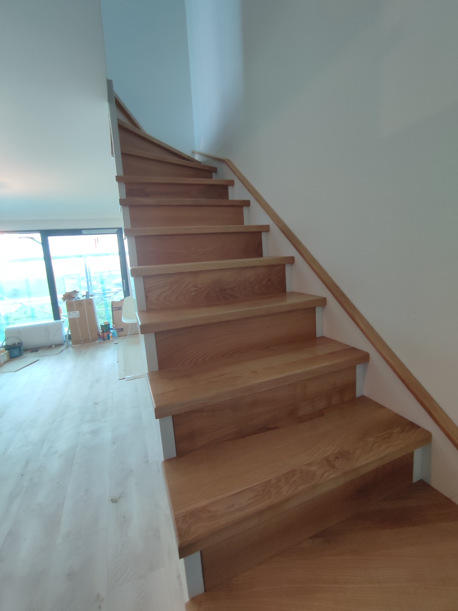 Nowoczesne schody drewniane z jasnego drewna, z białymi wspornikami, prowadzące w górę, widok z dołu, w tle okno i kartony po remoncie.