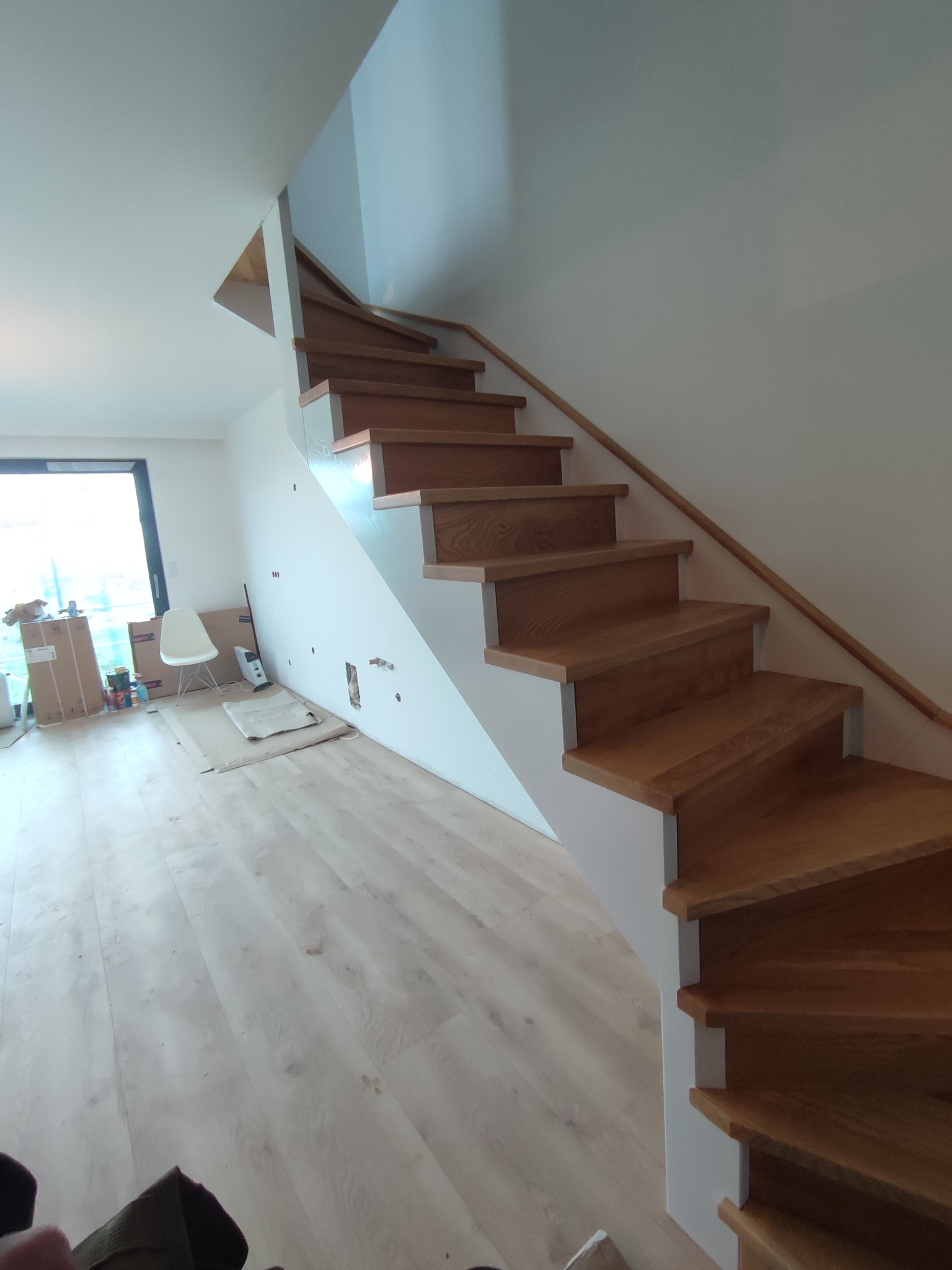 Nowoczesne schody drewniane z jasnego drewna, z białymi podstopnicami i prostą drewnianą balustradą, prowadzące na piętro w jasnym wnętrzu. Widoczne niedokończone prace wykończeniowe w tle.